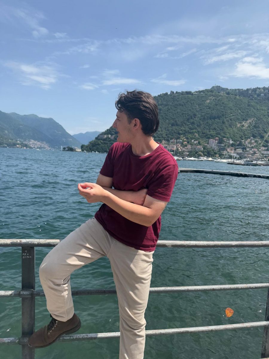 A student stands by a lakeside railing, gazing to the side. He wears a maroon shirt and beige pants with one foot propped up on the railing. Behind him are green hills, waterfront buildings, and a calm lake under a bright blue sky.