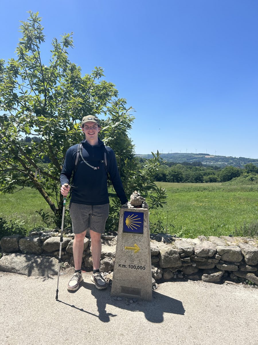 Person standing outdoors on a sunny day next to a Camino de Santiago milestone marker indicating 100 kilometers remaining. The individual is wearing a dark long-sleeve shirt, gray shorts, a green cap, and hiking shoes, holding a trekking pole. Behind them is a green field with trees and wind turbines visible in the distance under a clear blue sky.