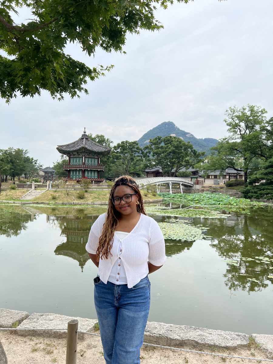 A UConn student stands in front of a serene pond with lotus leaves at the Hwangwonjeong Pavilion in Gyeongbokgung Palace, Seoul, South Korea. She is wearing glasses, a white top, and blue jeans, smiling gently with her hands behind her back. Traditional Korean architecture and a lush mountain are visible in the background under a cloudy sky, reflecting UConn's global engagement and cultural exchange.
