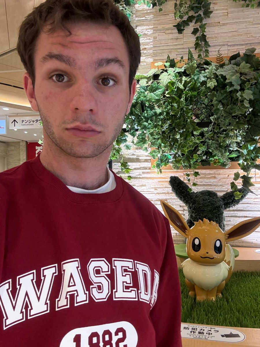 A student wearing a red WASEDA University sweatshirt takes a selfie indoors next to a decorative figure of Eevee, a popular Pokémon character. The Eevee statue is placed on artificial grass and partially surrounded by lush green plants and foliage decor, giving the area a garden-like feel. The setting appears to be inside a modern building with directional signage and tiled walls in the background.
