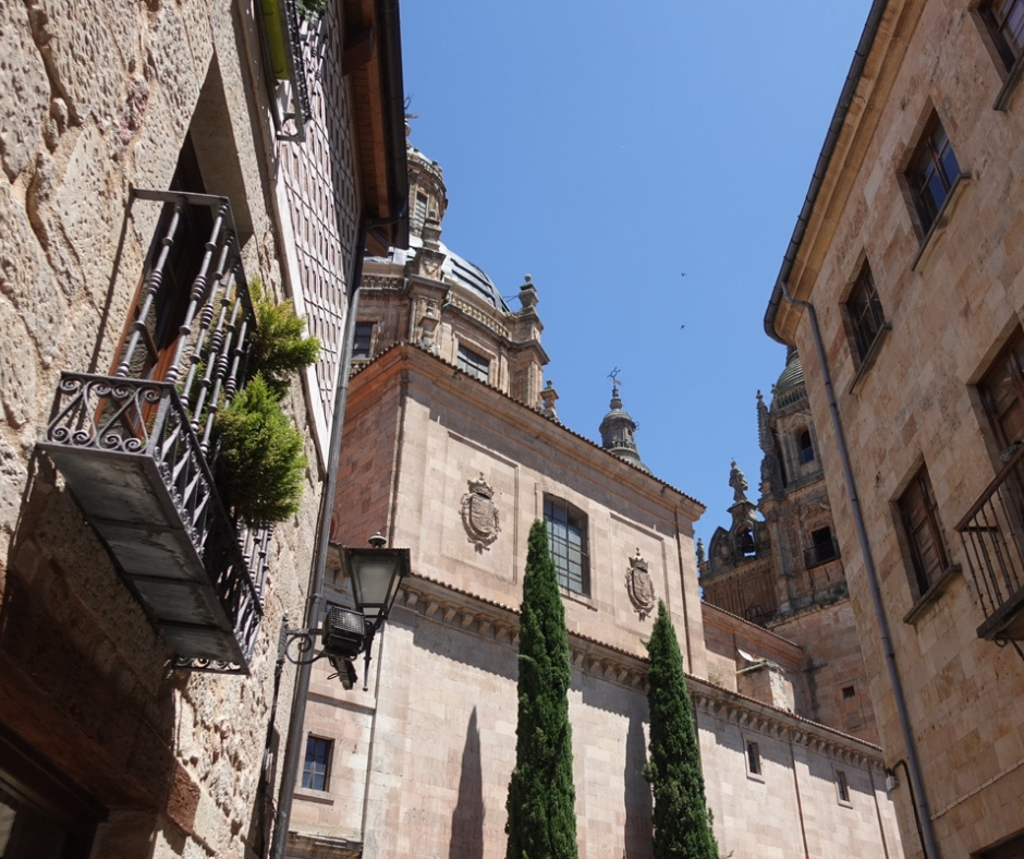 Historic sandstone buildings and a cathedral dome in Salamanca, Spain, viewed from a narrow street with wrought iron balconies and cypress trees. The architecture features ornate stone carvings and classical European design under a clear blue sky.