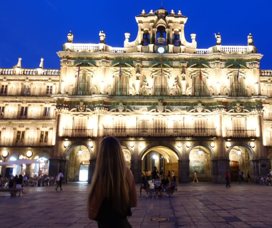 Evening view of a student standing in Plaza Mayor in Salamanca, Spain, facing the illuminated baroque-style City Hall building. The plaza is lively with people dining and walking under the deep blue sky, reflecting the vibrant local culture and student experience abroad