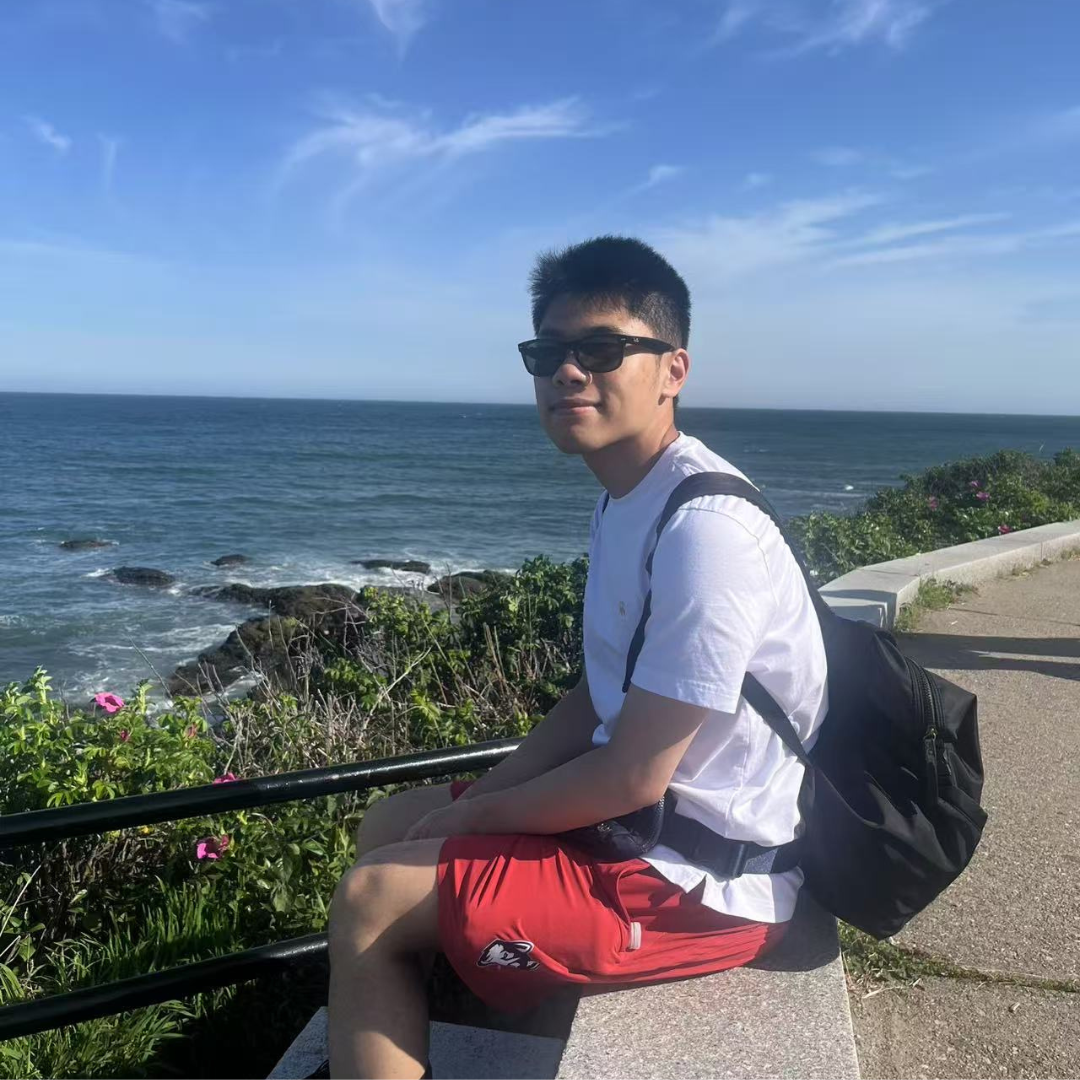 Photo of UConn student sitting by the ocean. He is wearing a white t shirt, red shorts, black backpack, and black sunglasses. His body is facing the ocean but he is facing the camera.