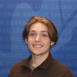Student is smiling at the camera. He has long blonde hair, and is wearing a brown quarter zip. He is posed in front of a blue backdrop, and the photo is shot shoulders up.