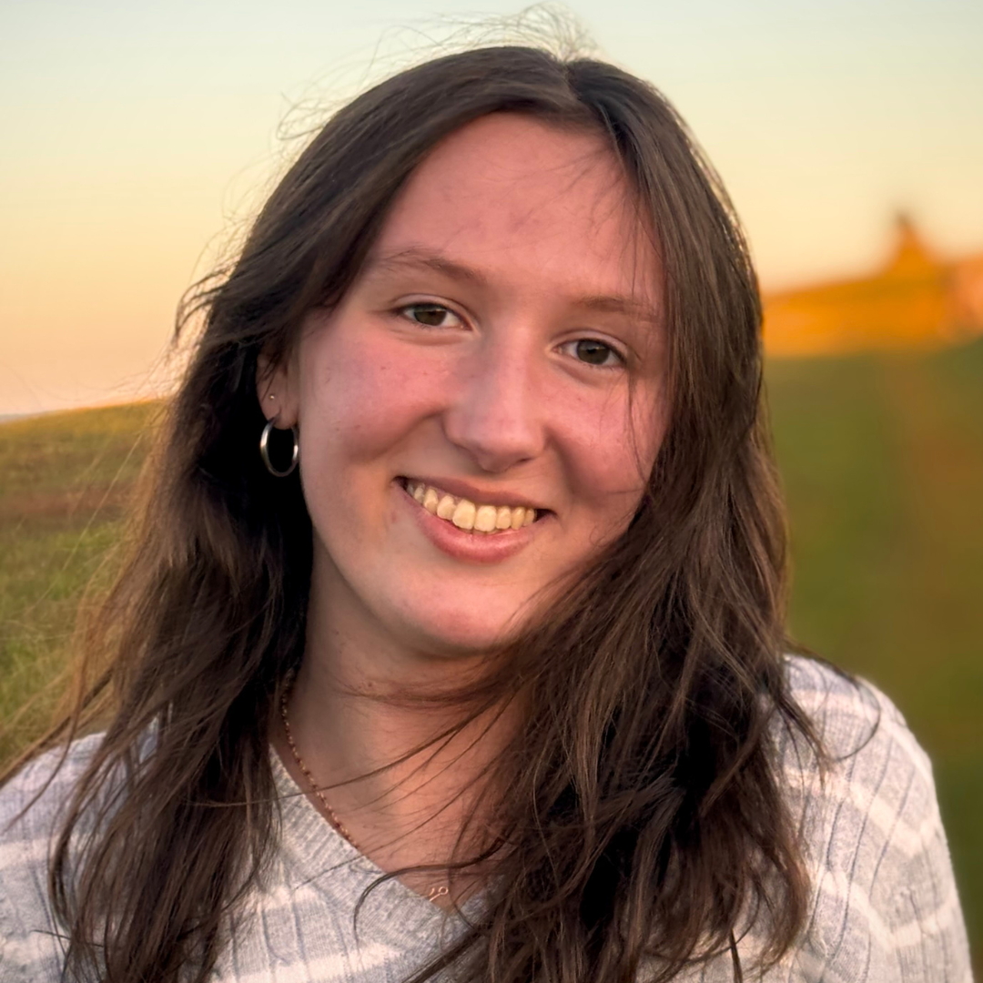 Headshot of UConn student smiling at the camera. She has short brown hair, is where a grey and white striped shirt, and is posed in front of a hill during sunset.