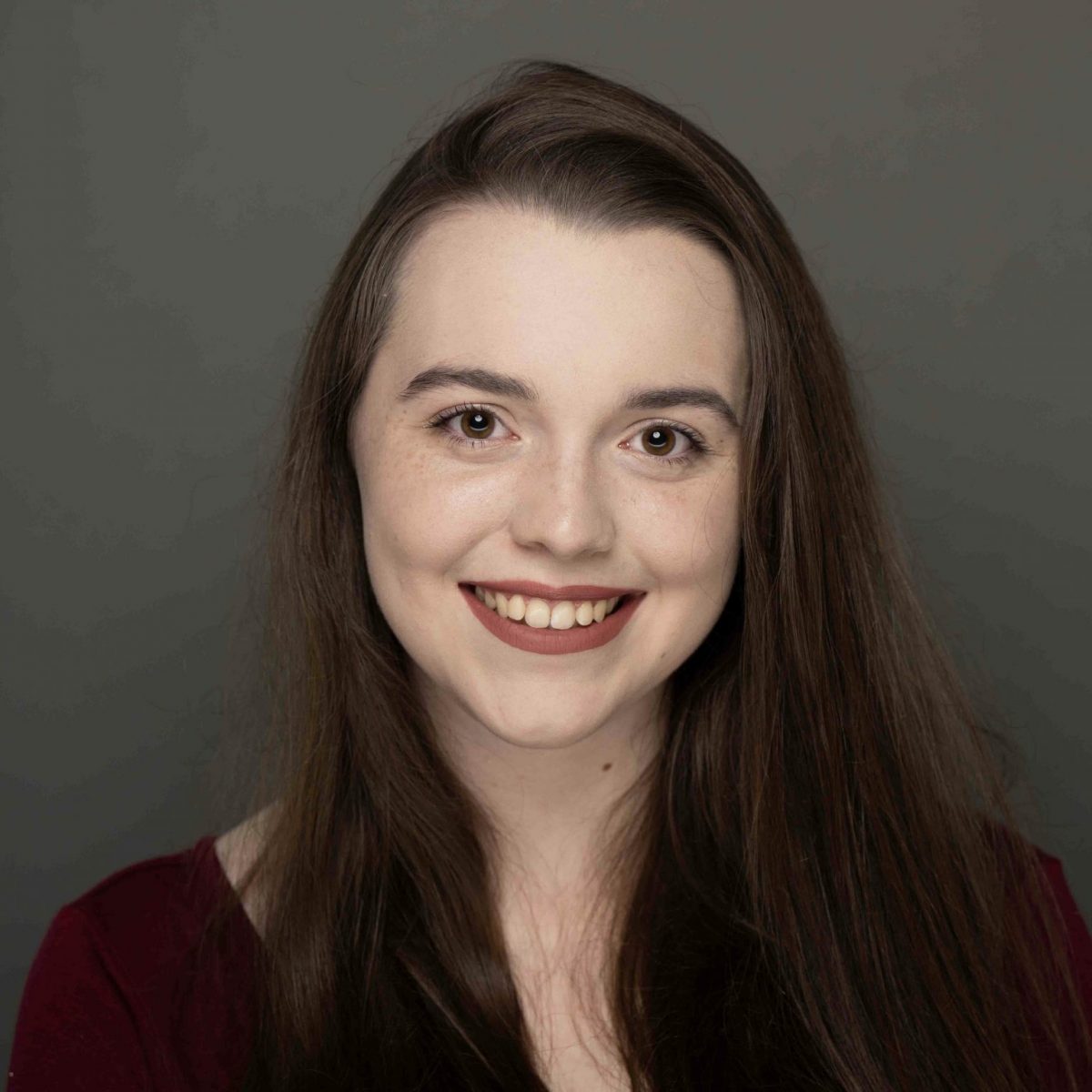 Student is smiling at the camera. She has long brown hair parted to the side, is wearing a maroon long sleeved top, and maroon lipstick. She is posed in front of a grey background.