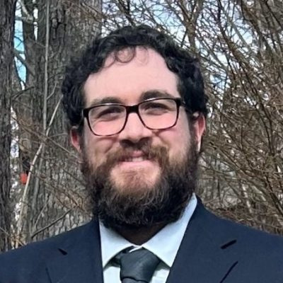 Student is smiling at the camera. He is wearing a navy blue suit and tie, glasses, and is posed outside with woods visible in the background.