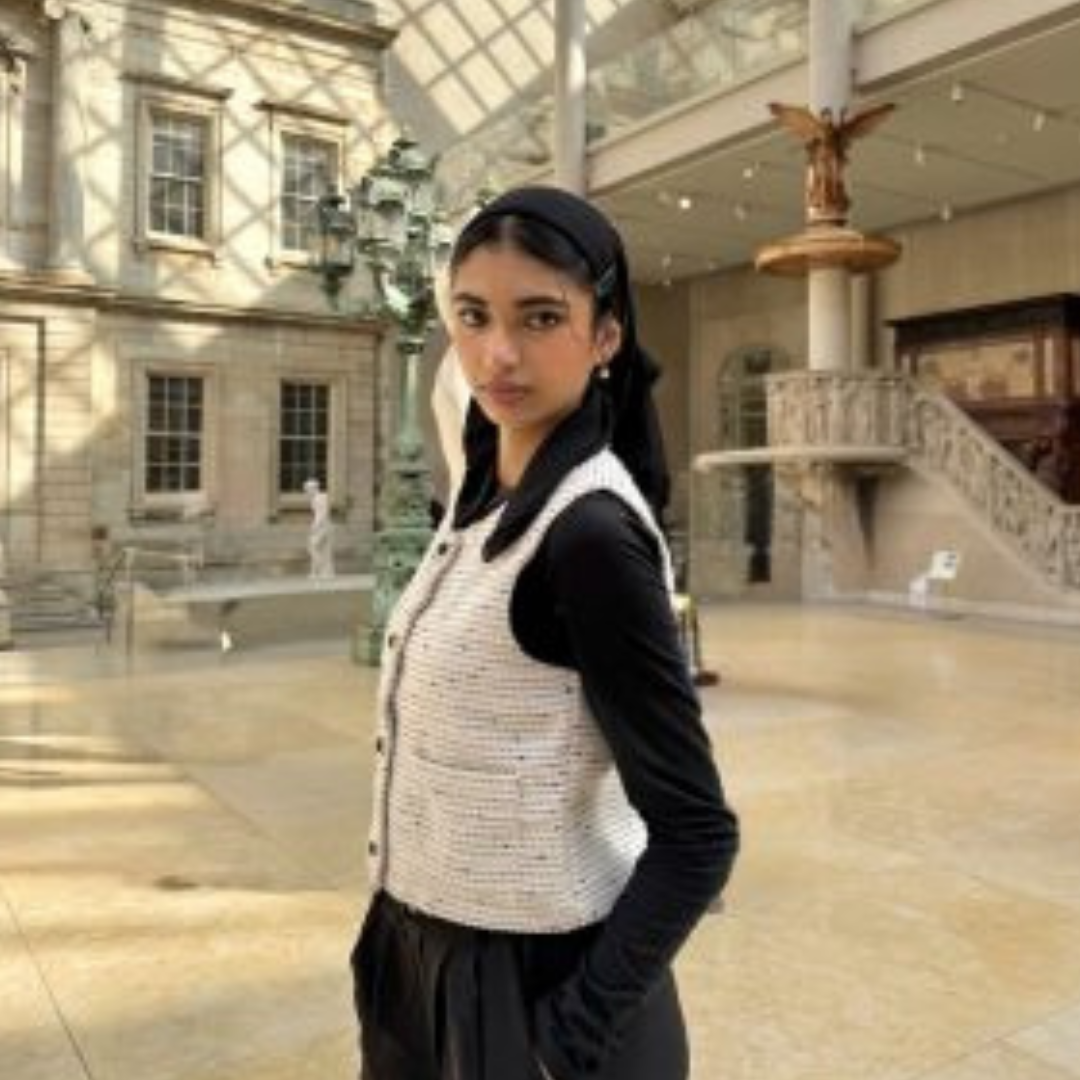 Student smiling at the camera in a long sleeve black dress, white vest, and black hijab. Student is posed with her hands in her pocket in front of an art museum background.