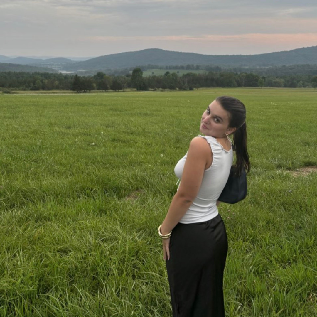 Photo of UConn student. She is wearing a white top, and black pants. She is posed looking over her shoulder in a green field with mountains on the horizon.
