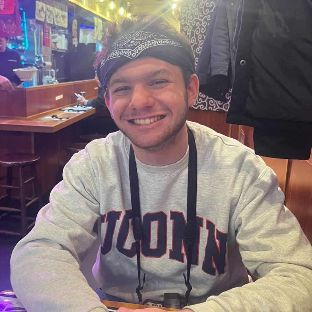 A man smiling at a table in a lively restaurant, wearing a UConn sweatshirt and a bandana.