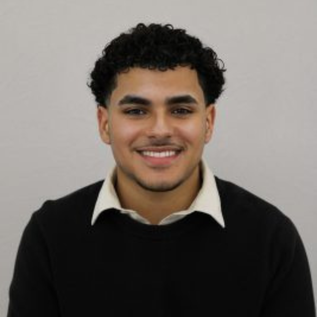 Student is smiling at the camera. He is wearing a white collar shirt underneath a black long sleeved sweater. He has black curly hair and is posed with both arms at his sides.