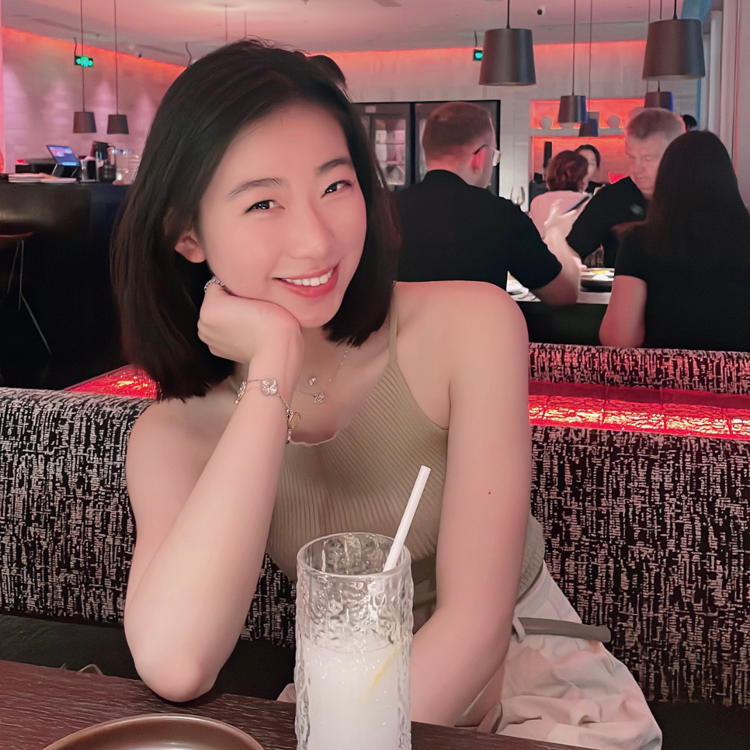 Photo of UConn student. She has short black hair, is wearing a beige top, and is posed at a restaurant with her face in her palm, and a drink in front of her.