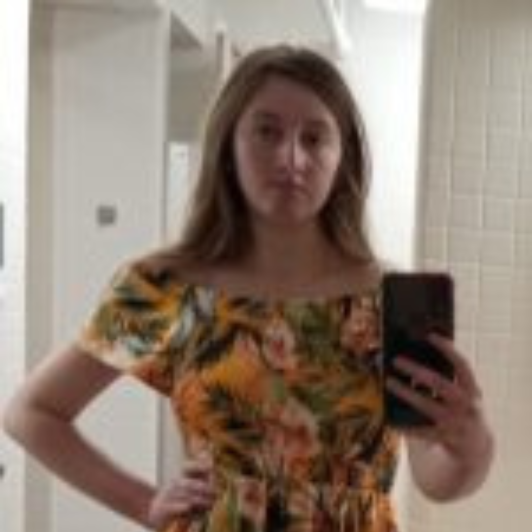 Student is smiling at the camera in the mirror. She is wearing a yellow and green floral dress, and has short brown hair. Student is posed with her hand on her hip in the mirror.