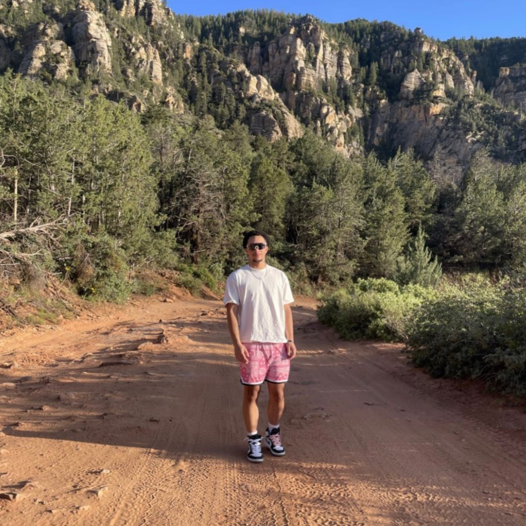 Student is smiling at the camera. He is wearing a white t-shirt, red shorts, black shoes, and black sunglasses. he is posed in front of a mountainous landscape.