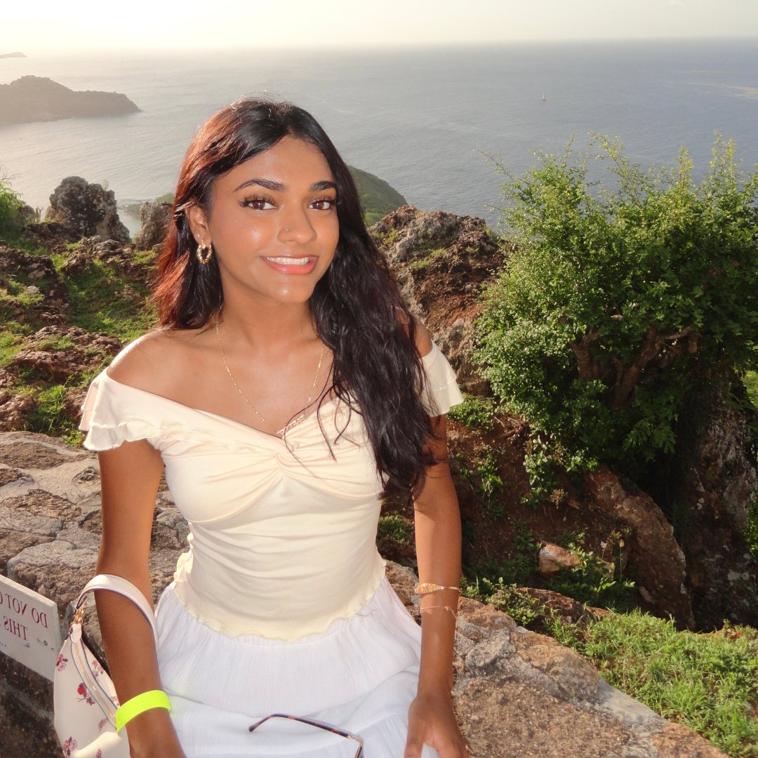 A woman smiling while sitting on a stone ledge with a scenic ocean and rocky landscape in the background.