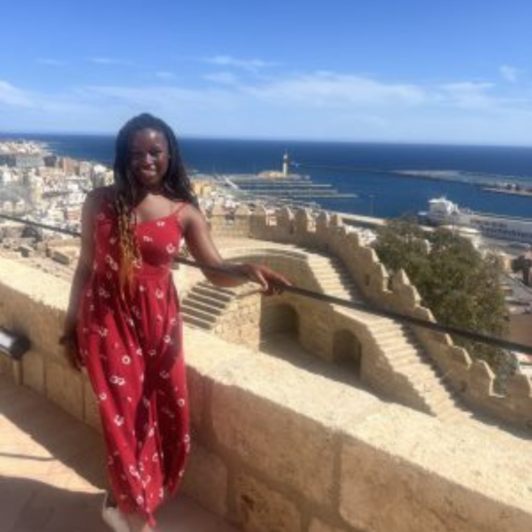 Student is smiling at the camera. She has black braids, is wearing a red dress and is posed in front of a view of the ocean at her study abroad destination.