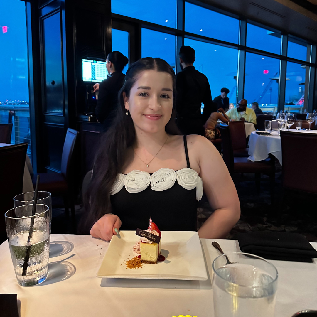Smiling woman in a black dress with white floral details, seated at a restaurant with dessert in front of her.