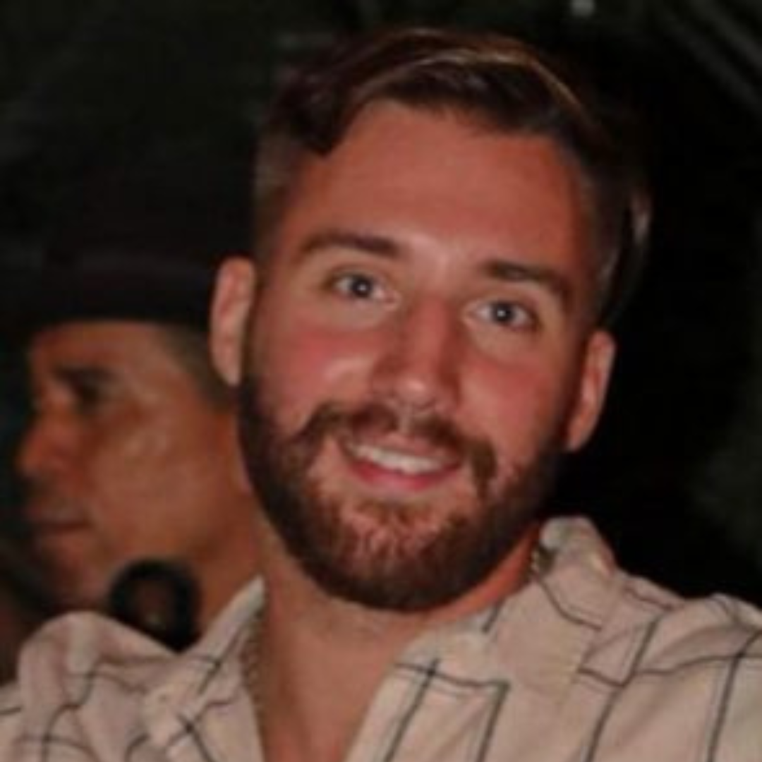 Student is smiling at the camera. He has a brown hair, and is wearing a cream and blue plaid shirt. The photo is shot shoulders up,