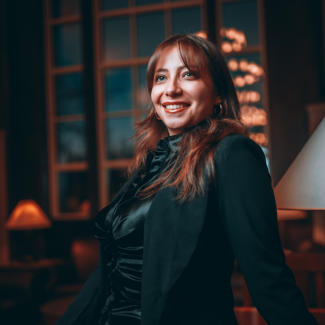 A woman smiling and posing indoors in a dimly lit, elegant setting, wearing a black outfit and blazer.
