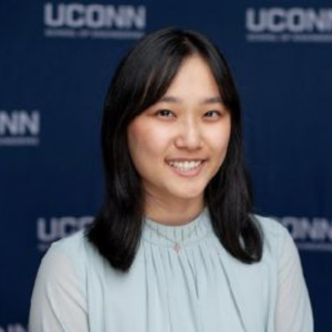 Student is smiling at the camera. She has short black hair and is wearing a sky blue colored blouse. She is posed in front of a navy UConn background.