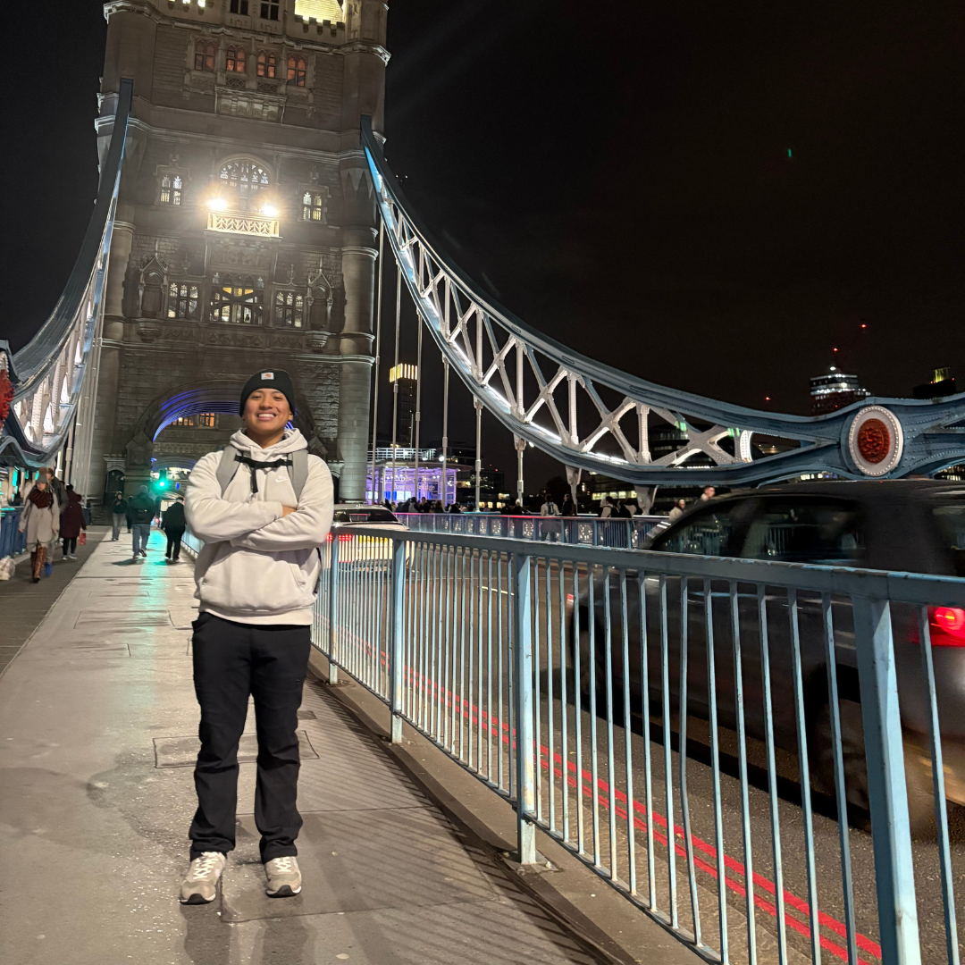 Student is standing in front of a bridge abroad, with his arms crossed. It is night time, and he is wearing a grey sweatshirt, black pants, beanie, and is smiling at the camera.