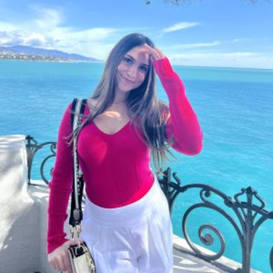 Student is smiling at the camera. She has long brown hair, is wearing a hot pink long sleeved shirt, and white pants. She is posed with her hand moving hair out of her face, in front of a bright blue ocean.