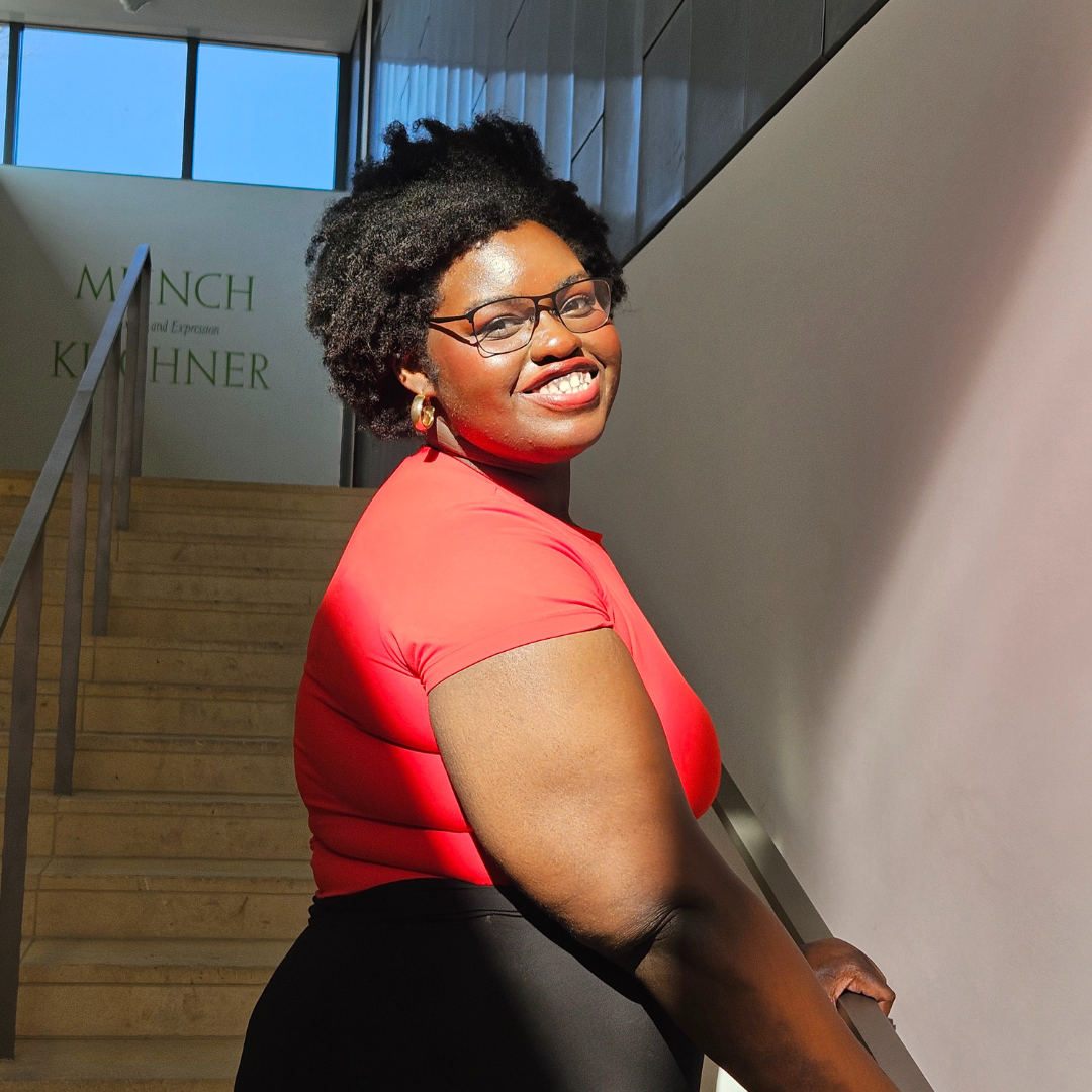 Student is smiling at the camera, and looking over her shoulder. She has short black hair, and is posed in front of a brightly lit staircase.