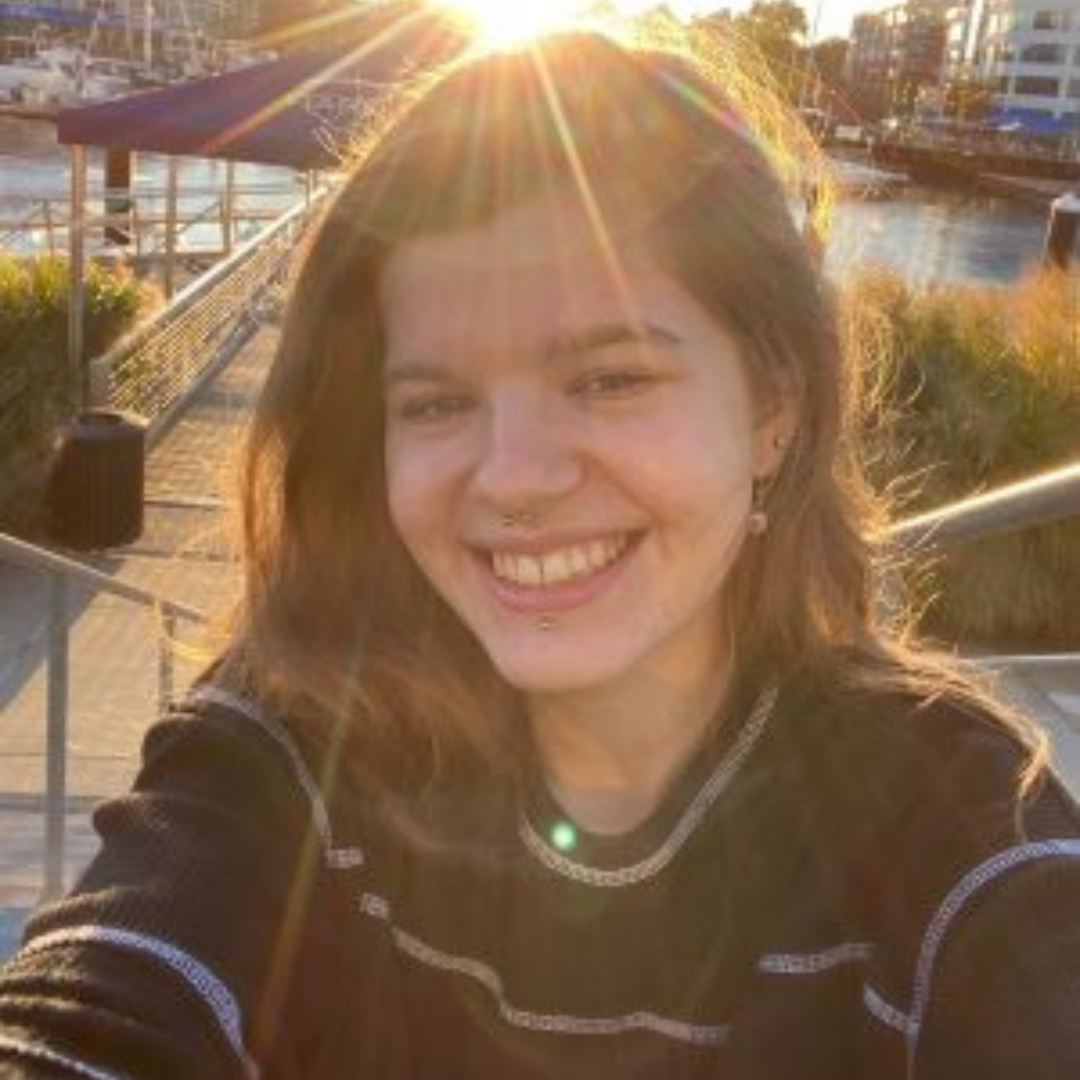 Student is smiling at the camera. She has short brown hair, and is wearing a black and white striped long sleeved shirt. She is posed in a city by the water, in front of the sun.