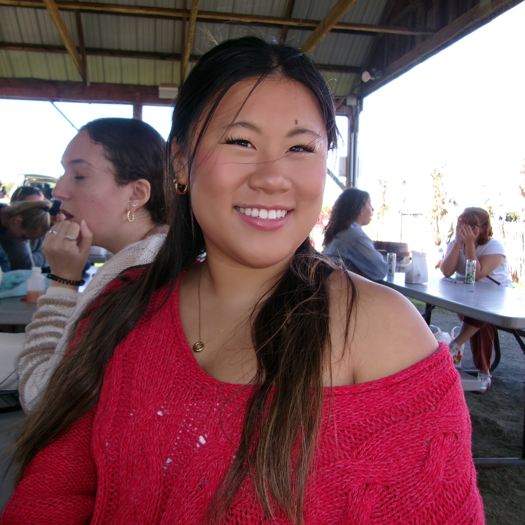 Student is smiling at the camera, posed under a gazebo. She is wearing a red sweater, has long brown hair in pigtails, and is posed in front of other people sitting at tables.