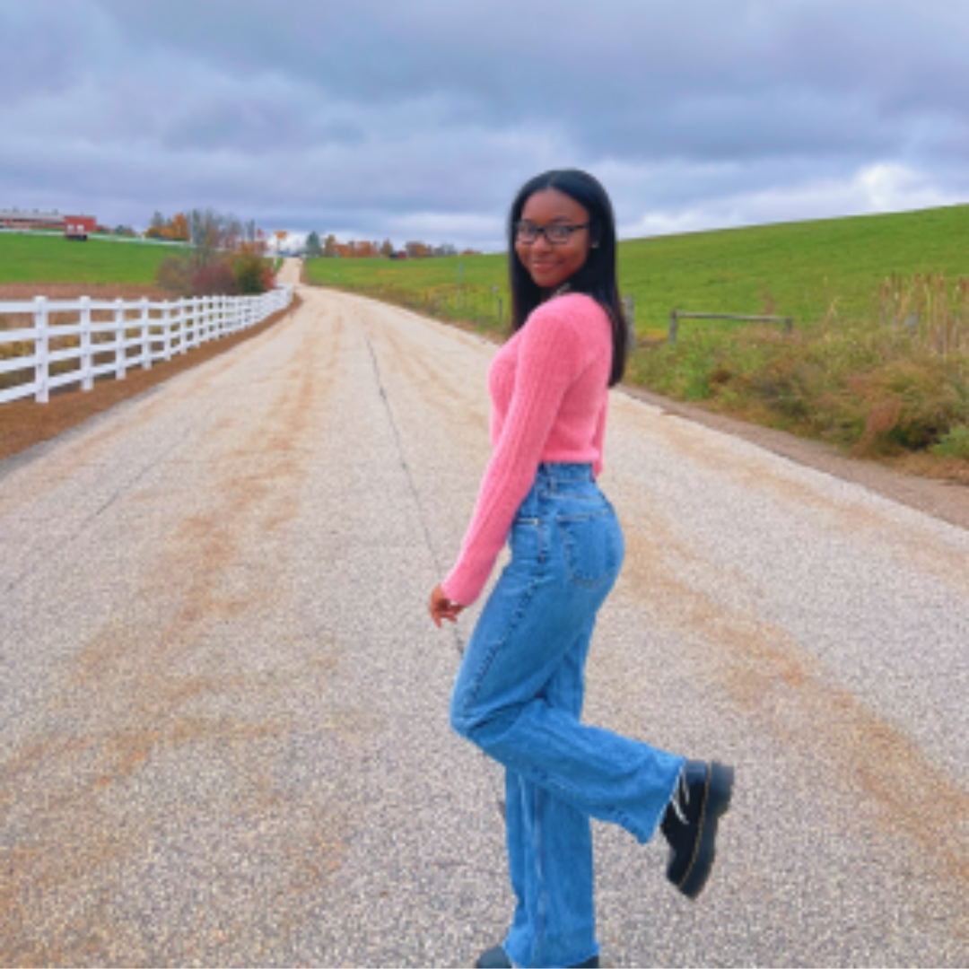 Student is smiling at the camera. She has medium length black hair, is wearing a pink long sleeved shirt and blue jeans. She is posed looking over her shoulder, wit her foot pointed up, in front of the road leading to Horsebarn Hill.