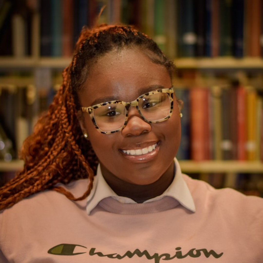 Student is smiling at the camera with printed glasses, red braids in a pony tail, and a white collared shirt underneath a pink Champion sweatshirt. She is posed in front of a shelf of books.