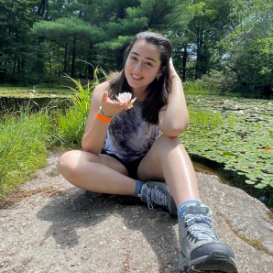 Student is smiling at the camera. She has short black hair, wearing a tie dye shirt, shorts, and sneakers. She is posed sitting down on a rock by a pond, holding a flower.