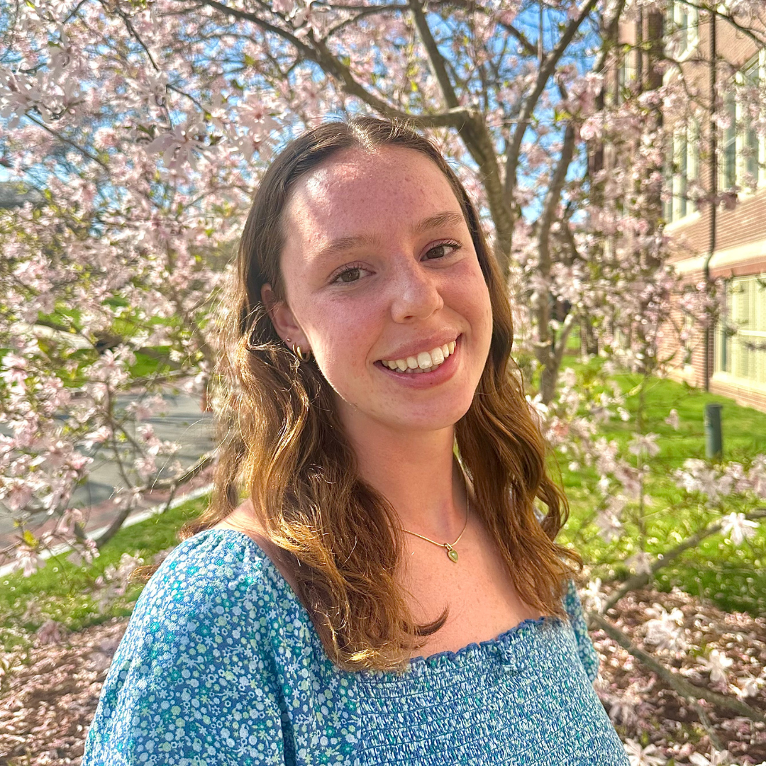Student is smiling at the camera in front of a tree of pink flowers. She has short wavy blonde hair, and is wearing a blue blouse.