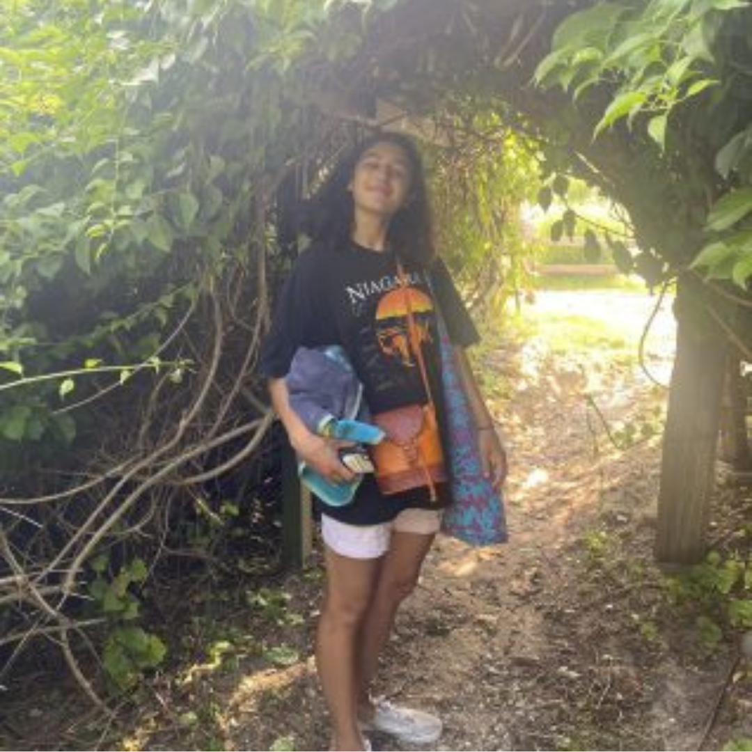 Student is smiling at the camera. She has short black hair, is wearing a baggy t-shirt, shorts, and sneakers. She is posed holding her tote bag under a tree in the woods.