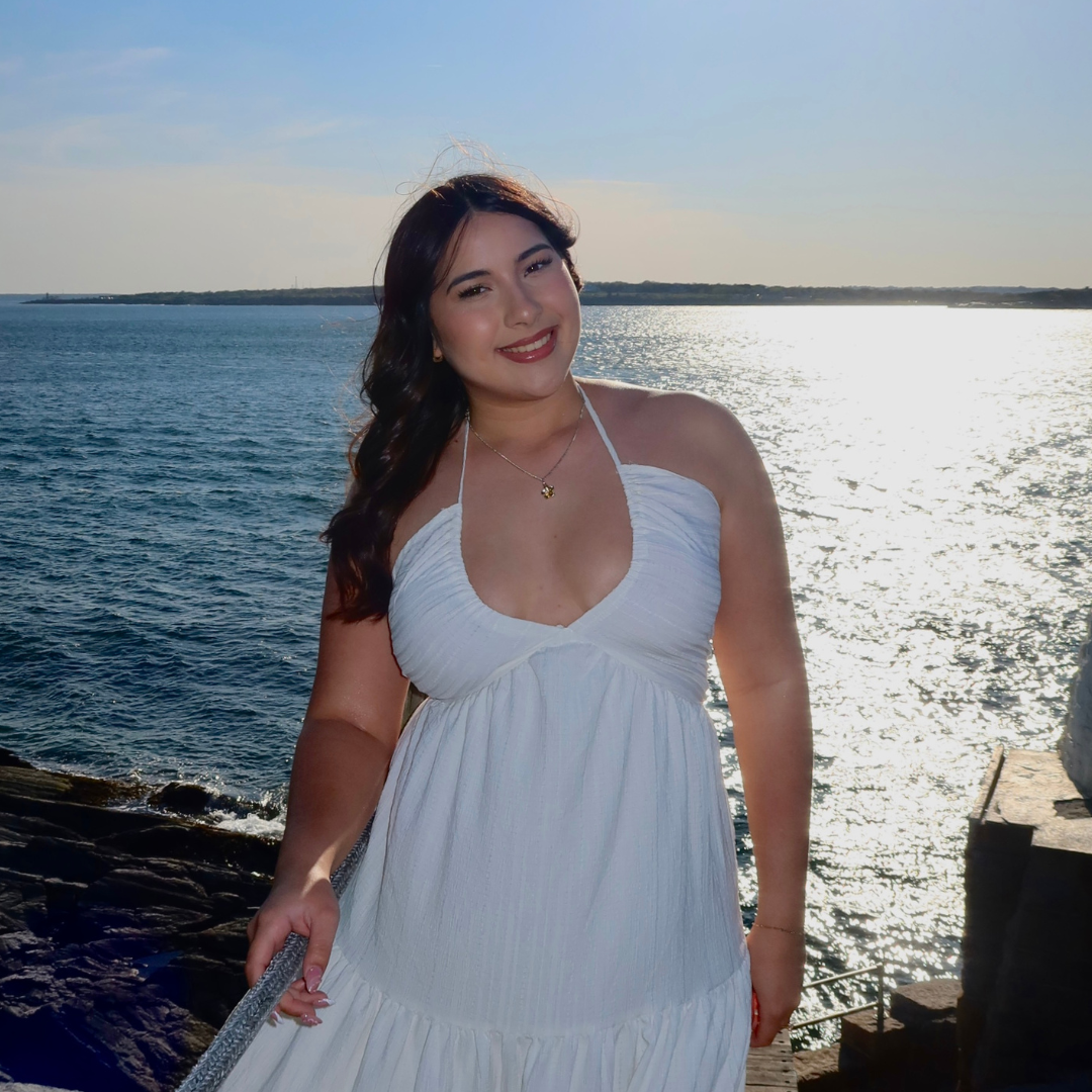 Student is smiling at the camera in a white dress. She is posed in front of an ocean background, with her hands at her sides.