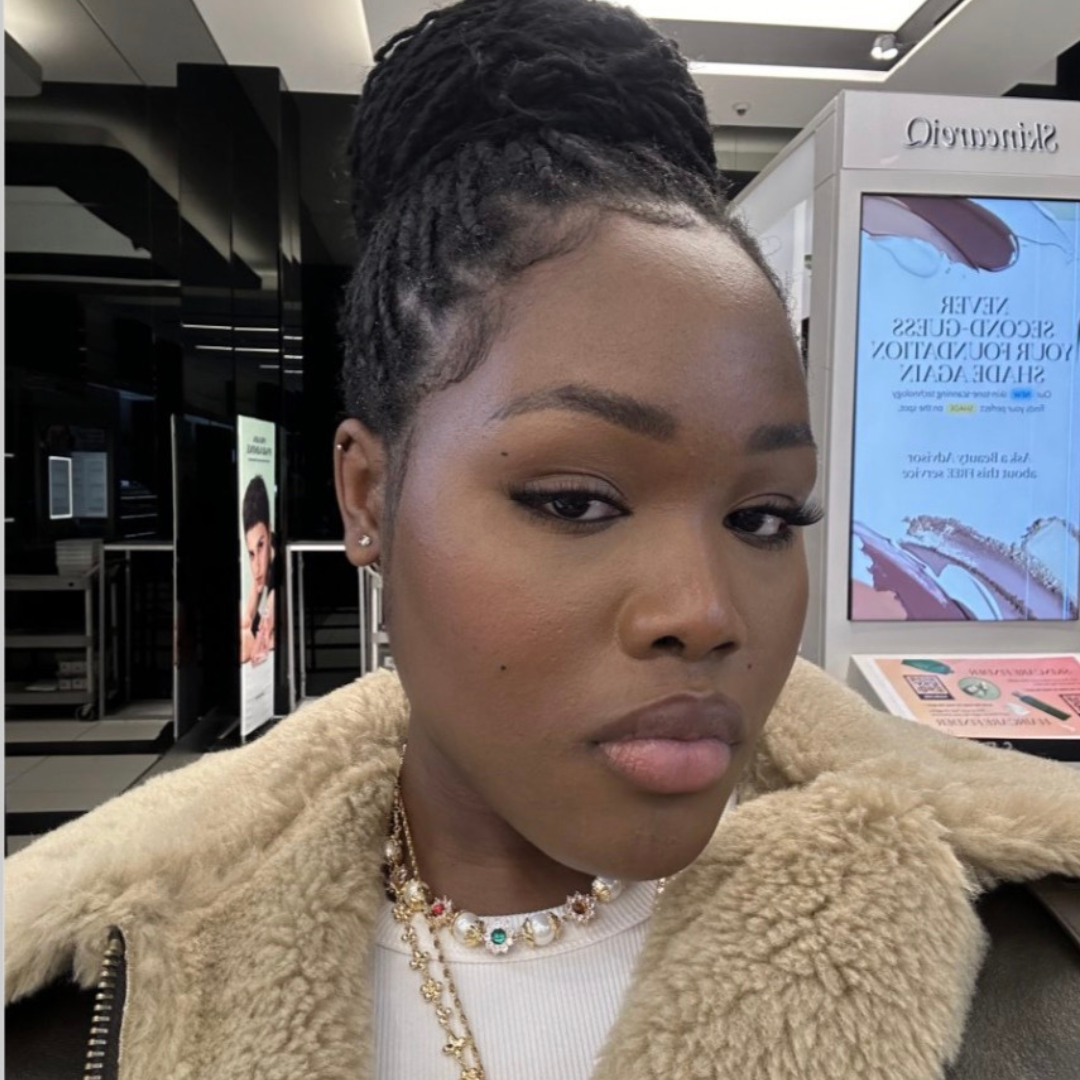 Student smiling at the camera. She is wearing a white shirt with a tan fur coat, and she has black braids tied into a bun. She is posed inside of a retail store.