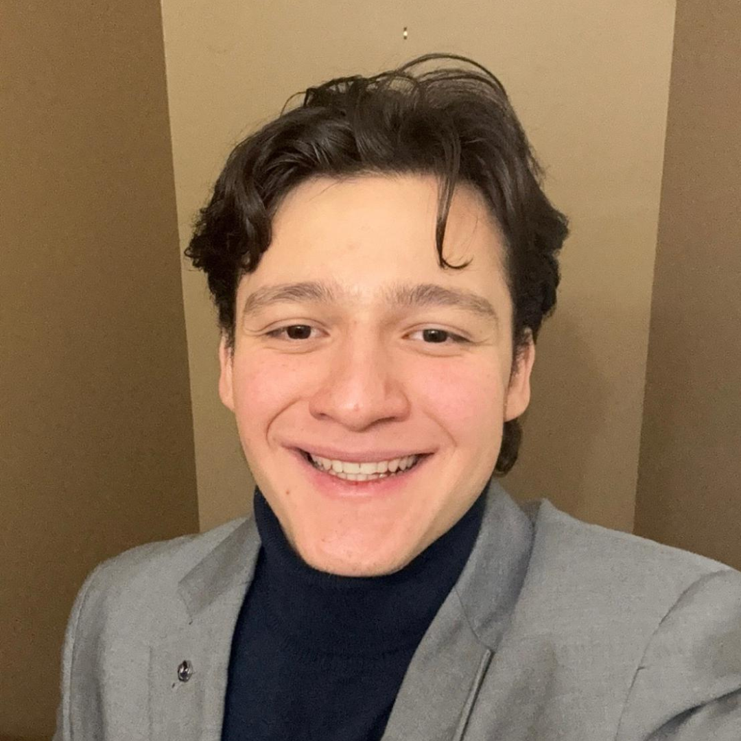 Smiling man with dark hair wearing a gray blazer and dark turtleneck, posed against a beige background.