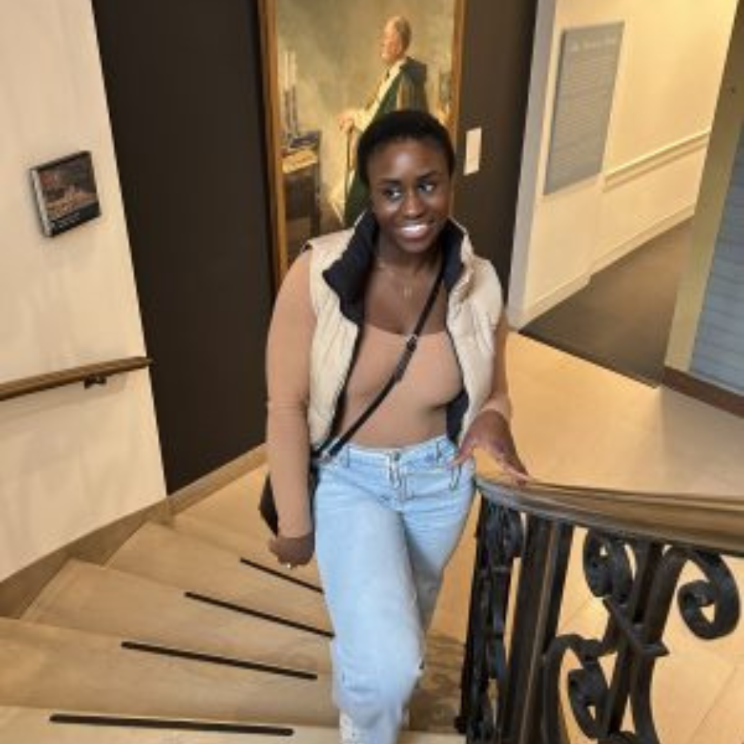 Student is smiling at the camera. She has short black hair, is wearing a tan tank top and vest, and blue jeans. She is posed on the bottom of a staircase inside an art museum.