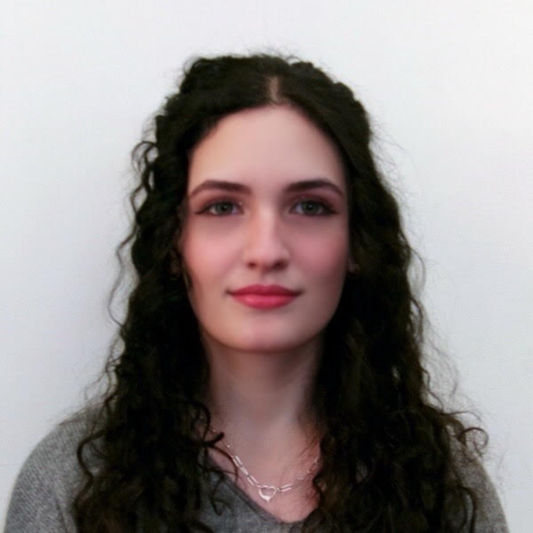 Student is smiling in front of a white background. She has long brown curly hair, and is wearing a grey top.
