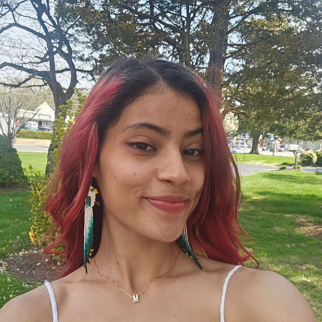 Student is smiling at the camera, outside in front of a tree filled background. She has short pink hair, and is wearing colorful feather earrings, and a white tank top.