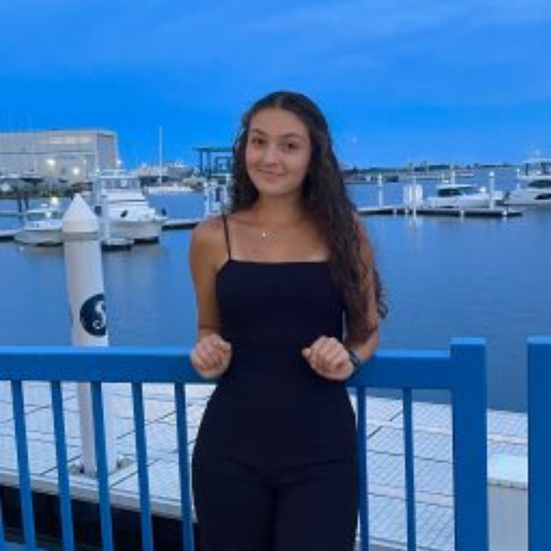 Student is smiling at the camera. She has long brown curly hair, and is wearing a black dress. She is posed in front of a marina of boats.