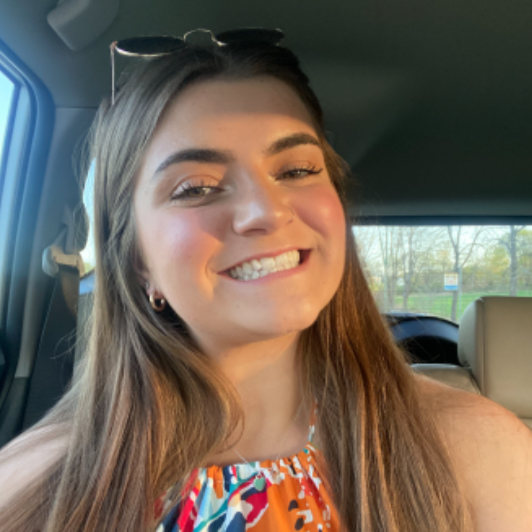 Student is smiling at the camera. She has long blonde hair, sunglasses on her head, and a colorful halter neck dress. The photo is a selfie of her inside a car.