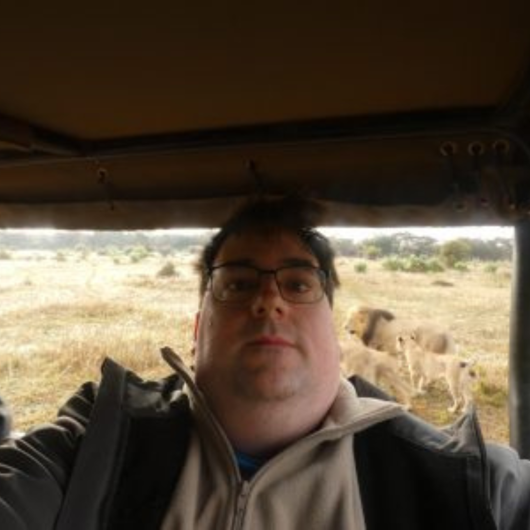 Student is smiling at the camera. He has black hair and is wearing a cream colored quarter zip and grey jacket. The photo is a selfie on a safari from a vehicle with animals shown in the background behind him.