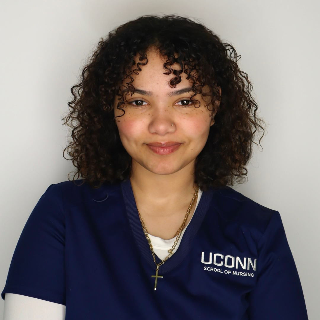 Student is smiling at the camera with her hands down. She has short brown curly hair, is wearing a white long sleeve, and a UConn School of Nursing shirt on top. She is posed in front of a plain background.