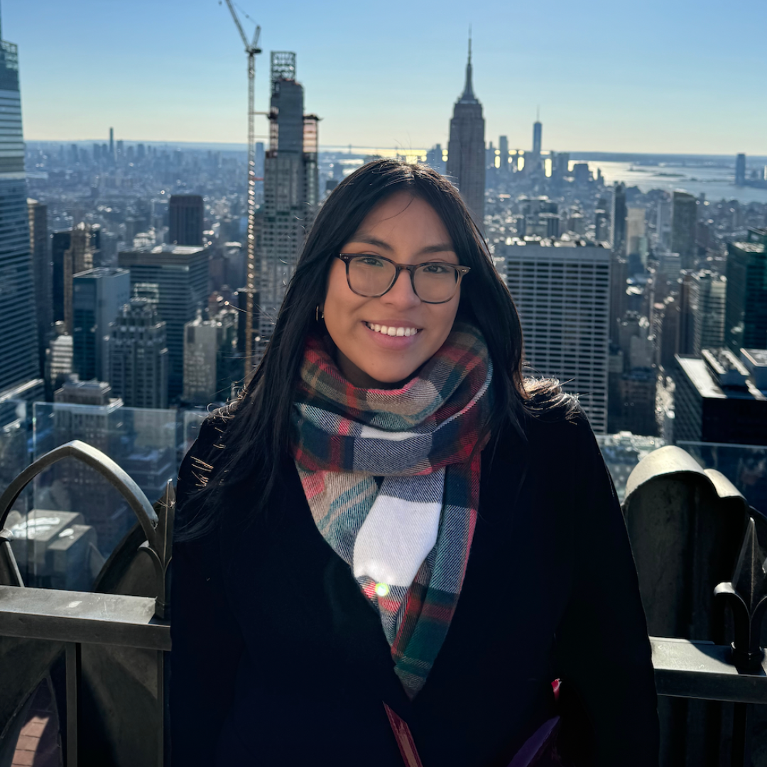 Student posed smiling at the camera with long black hair, black glasses, a black winter coat, and a plaid scarf. Student is posed infront of the New York City Skyline.
