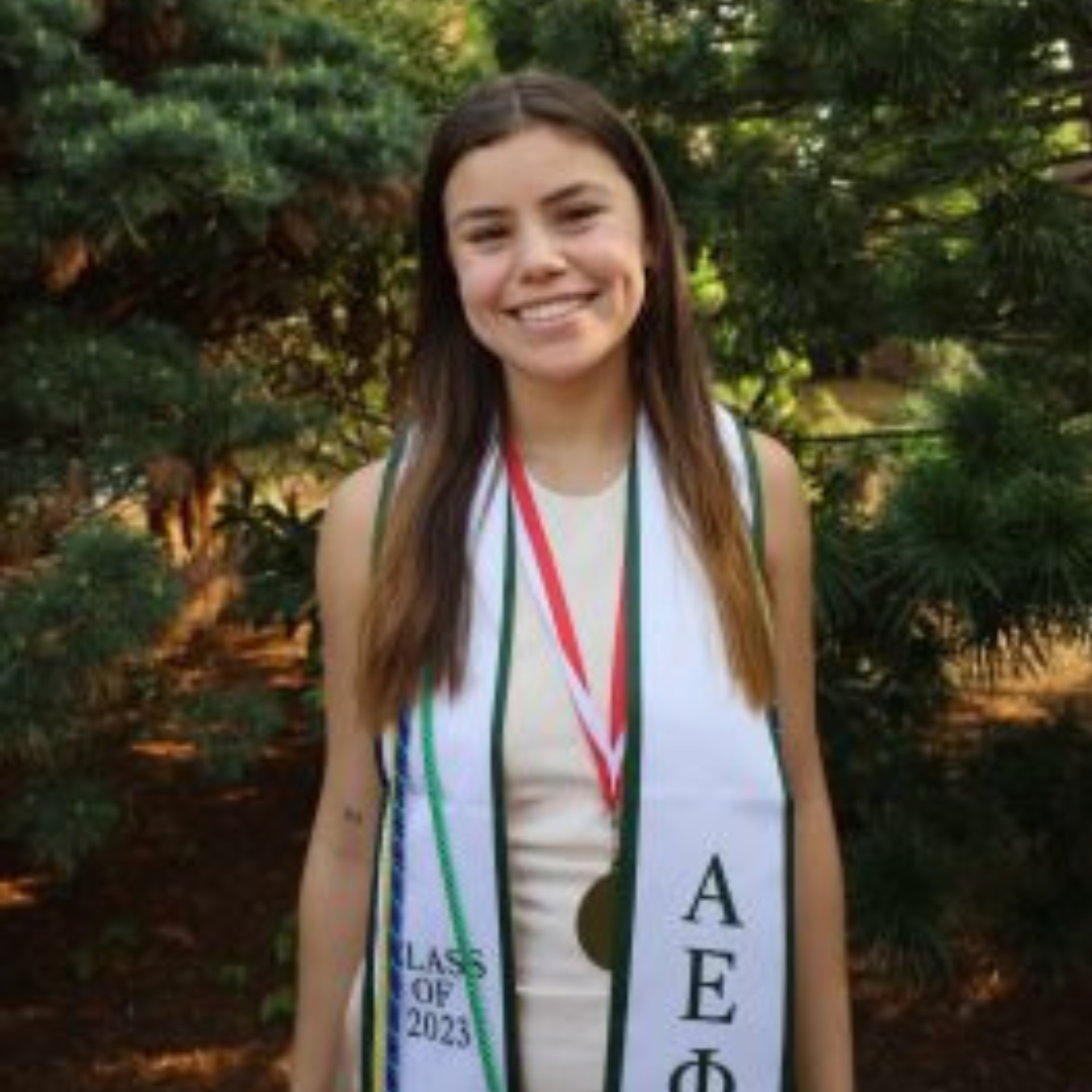 Student is smiling at the camera. She has long brown hair, is wearing a white dress, and her graduation medals/tassels. She is posed in front of trees on the Storrs campus.