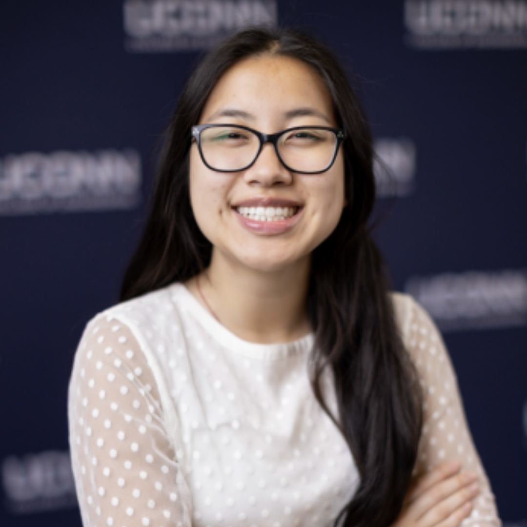 Student smiling at the camera with black glasses, straight black hair, and a white long sleeved blouse. She is posed in front of a blurred background that says "UConn." She is posed with her arms crossed, and her hair half in front.