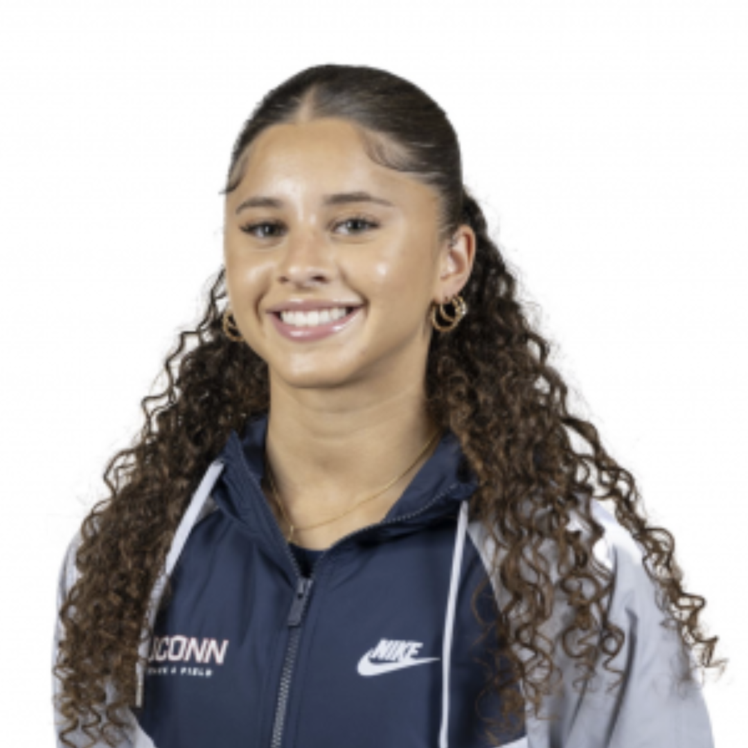 Student smiling in front of a white background. She has brown curly hair, and is wearing a UConn x Nike zip-up.