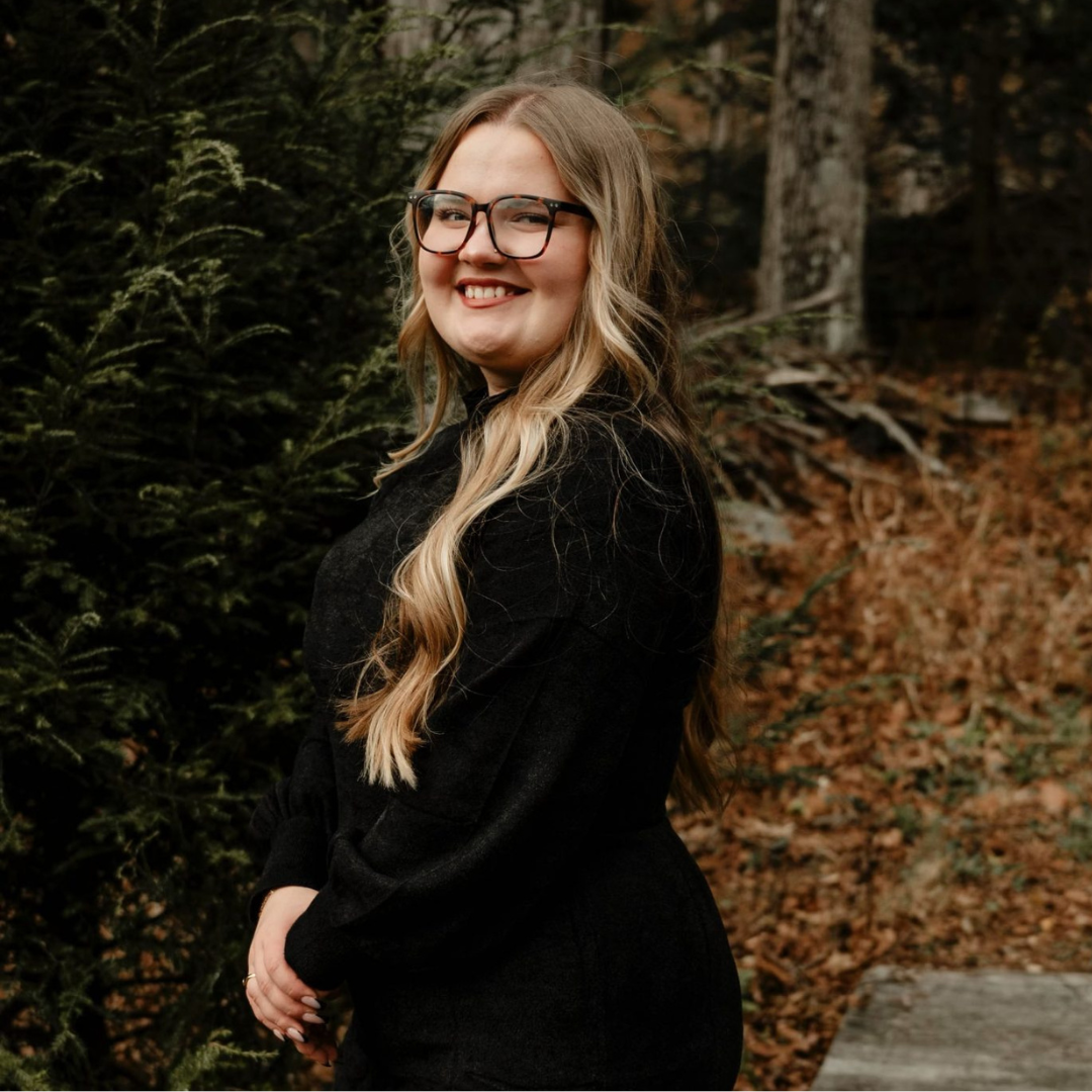 Student is smiling at the camera. She is wearing black glasses, a black long sleeve top, and she has long blonde wavy hair. Student is posed outside in the woods with leaves behind her.