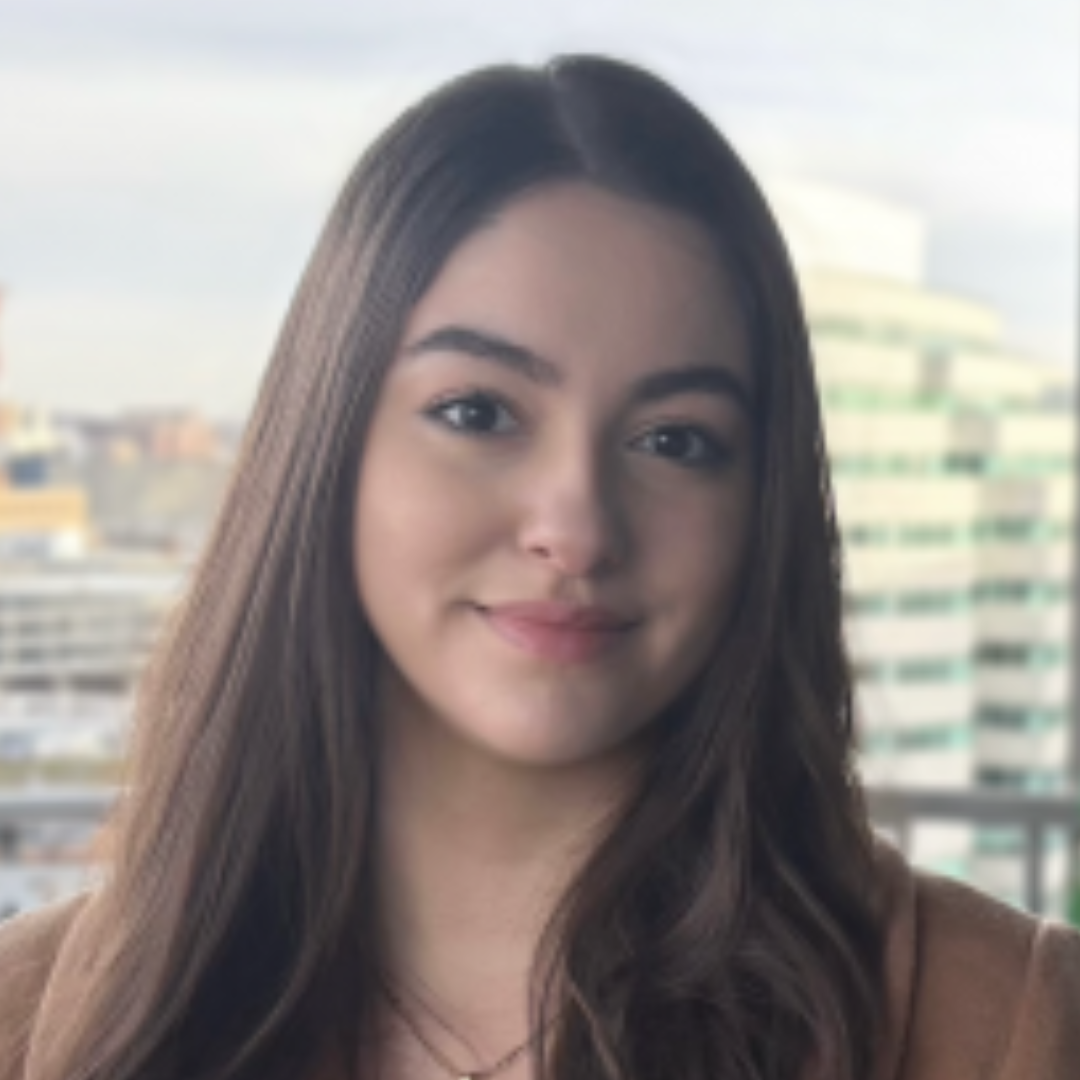 Student is smiling at the camera. She has long brown hair and is wearing a light brown blazer and necklace. She is posed in front of a window with a daytime city view behind her.